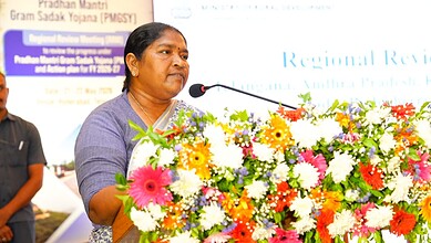 A woman speaking at a podium decorated with colourful flowers during a formal event in India.