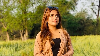 Sunlit woman with long brown hair standing in a lush green park during daytime.