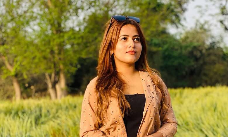 Sunlit woman with long brown hair standing in a lush green park during daytime.