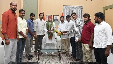 Group of men presenting a bouquet to an elderly man in a room with framed pictures and a window with curt.