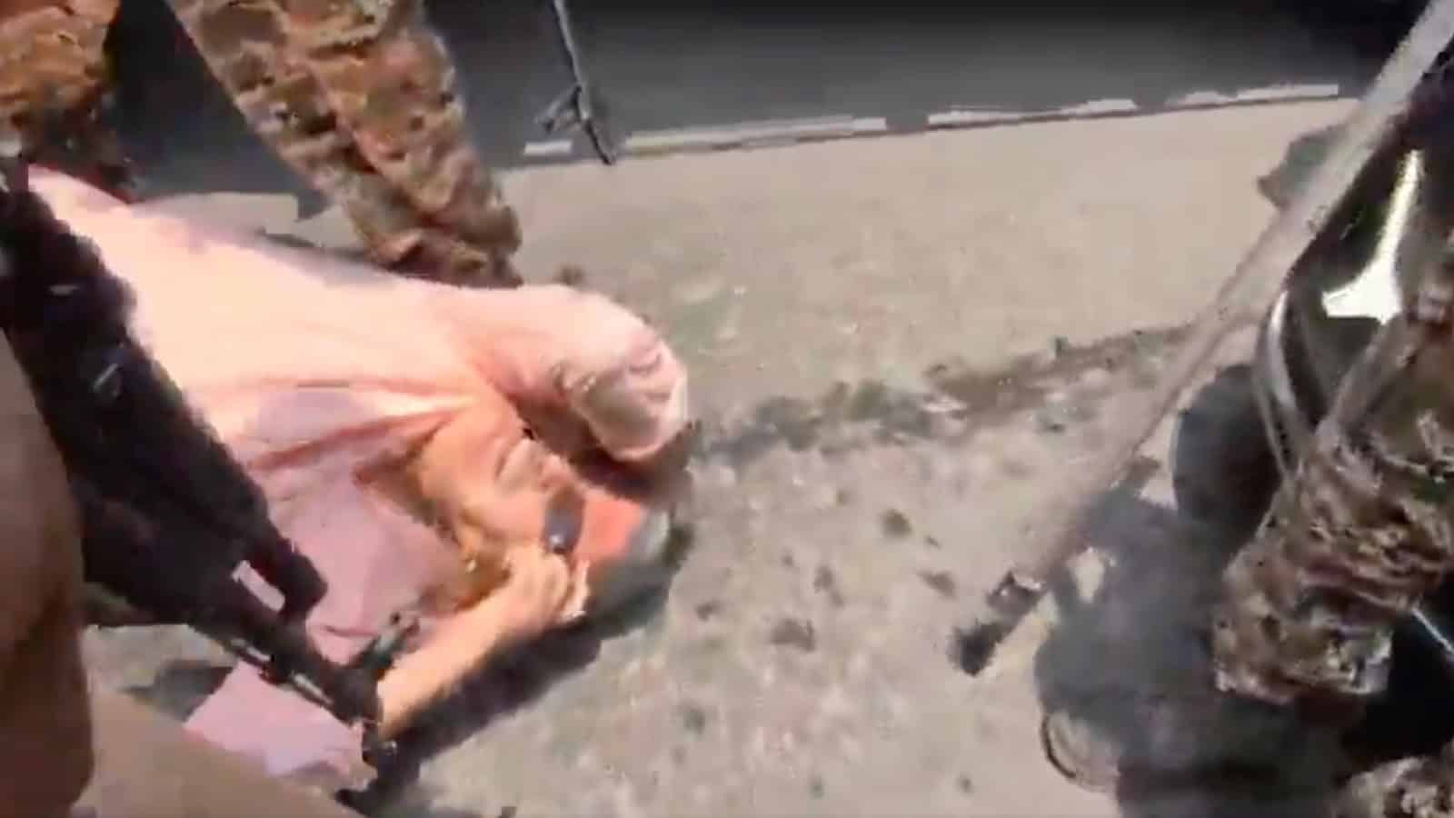A woman lying on the ground with military personnel around her, near a damaged vehicle on a street.