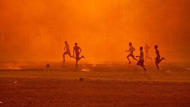 Children playing football on a field during sunset with an orange sky in the background.