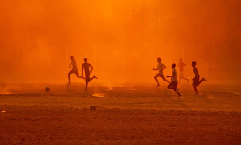 Children playing football on a field during sunset with an orange sky in the background.