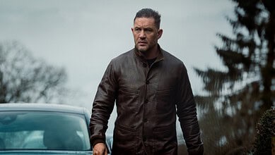 A man wearing a brown leather jacket standing outdoors with trees and cloudy sky in the background, captu.