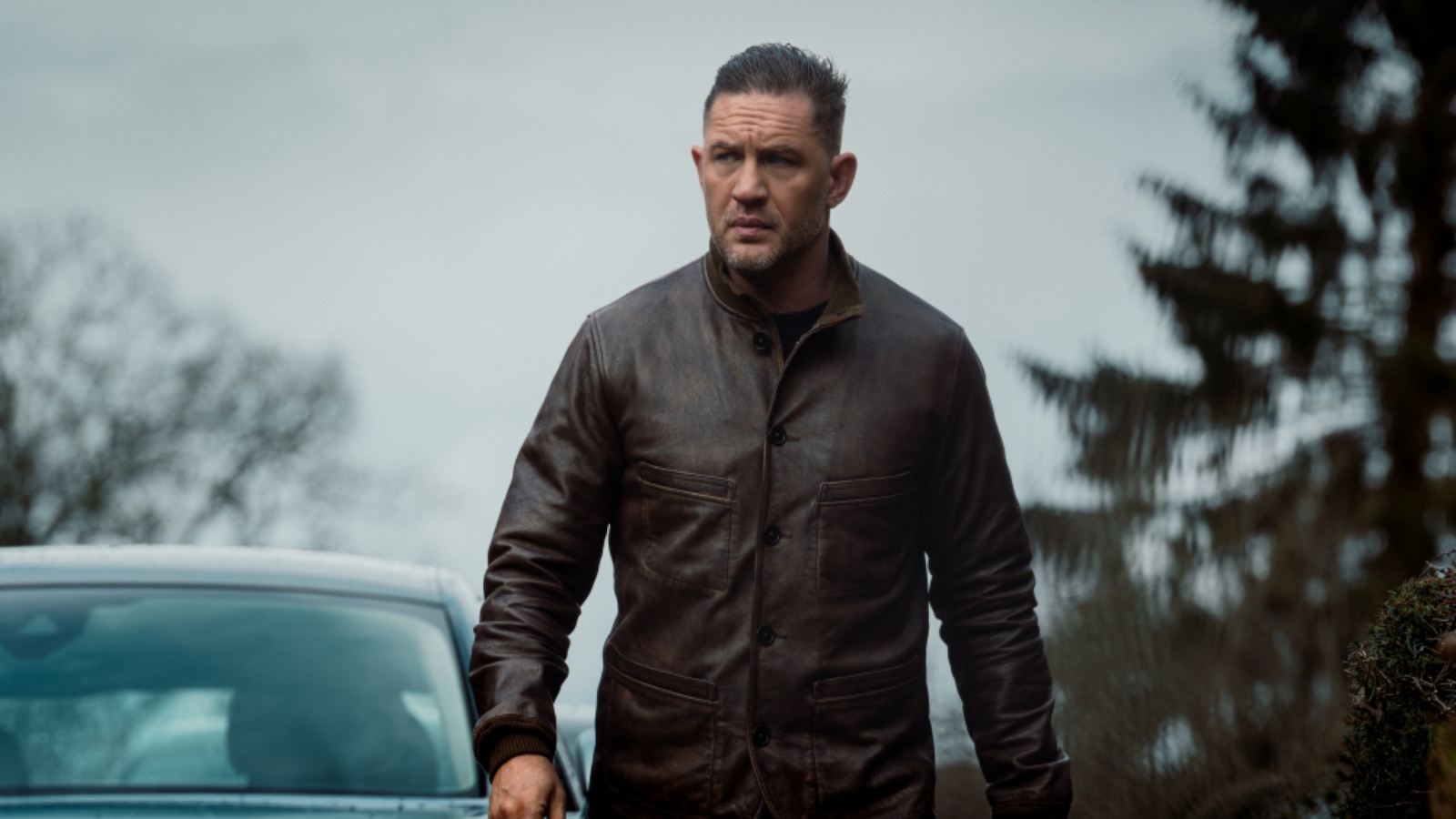 A man wearing a brown leather jacket standing outdoors with trees and cloudy sky in the background, captu.