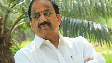 Close-up of an Indian politician with glasses and a white shirt, standing outdoors with greenery in the b.