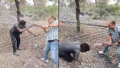 Two men fighting with sticks in a wooded area, one man in a checkered shirt and sunglasses, the other in.