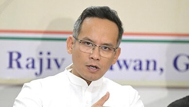 Politician speaking at a meeting about Assam election performance, wearing glasses and white attire.