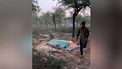Man walking past a makeshift shelter on fire in a rural landscape with trees and dry ground.