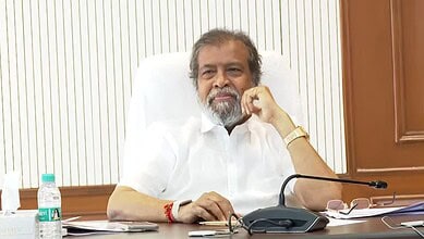 A man with grey hair and beard sitting at a desk in an office setting, wearing a white shirt and a gold w.