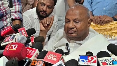 Senior man speaking to the media surrounded by microphones and journalists.
