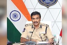 Indian police officer in uniform speaking at a desk with the Indian flag and a badge in the background.