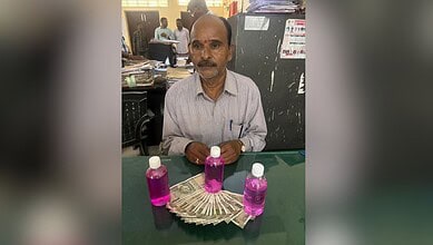 A man with a mustache sitting at a desk with three bottles of pink liquid and a fan of currency notes in.