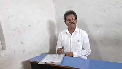 A person smiling and sitting at a desk with papers, wearing glasses and a white shirt.