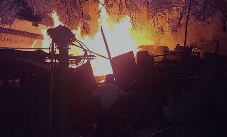 A firefighter battling a large blaze with intense flames and smoke in the background.
