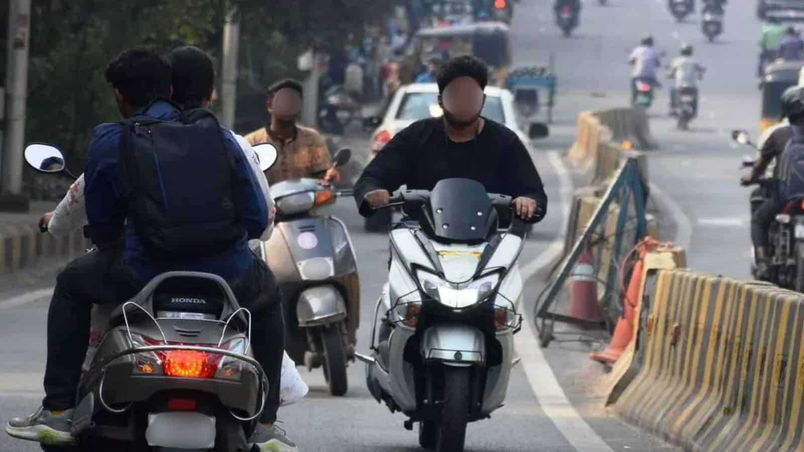 Scooter riders and pedestrians on busy urban street with traffic and road barriers in the background.