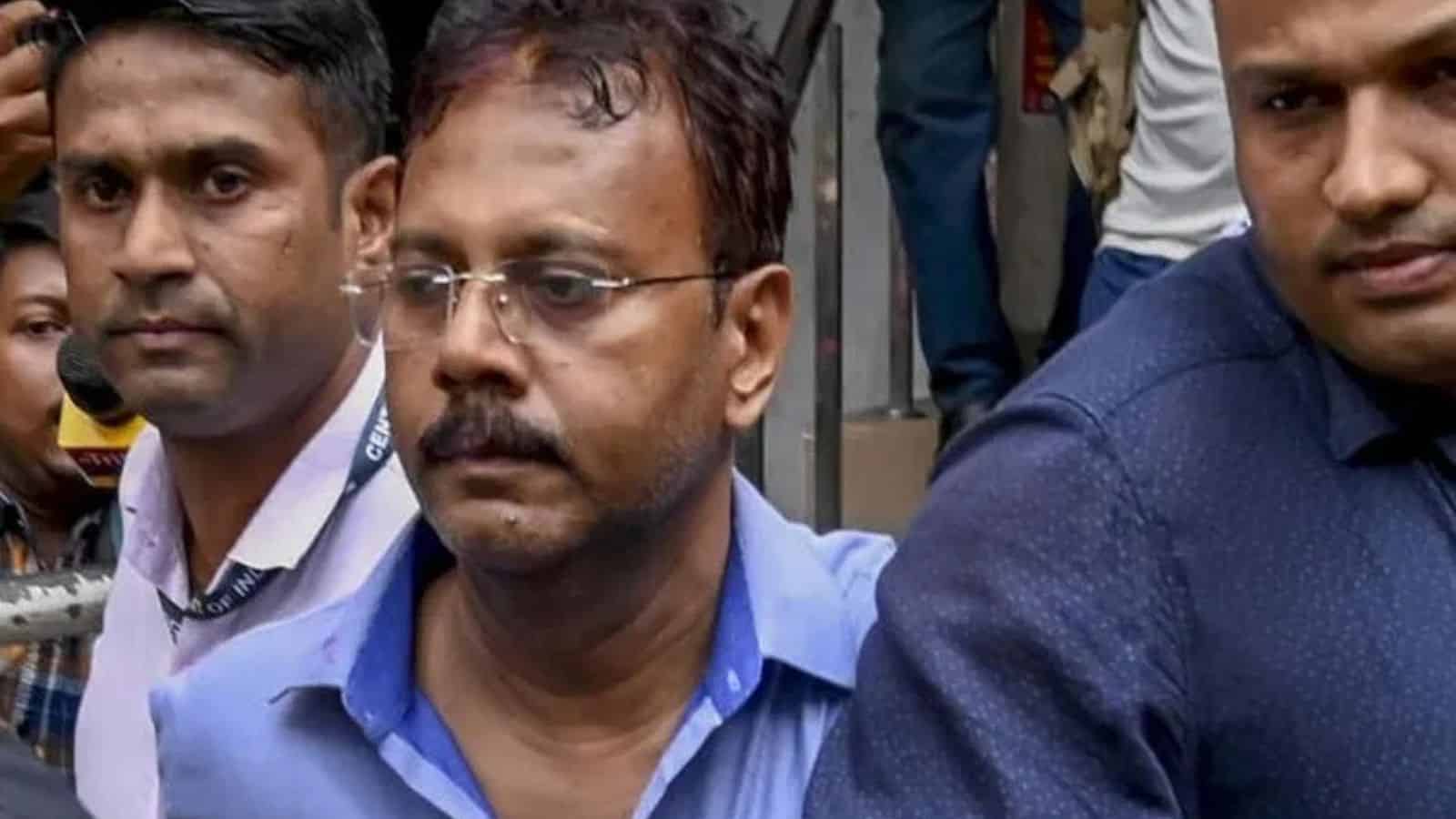 A man with glasses and a mustache in a blue shirt among a group of people in an outdoor setting.