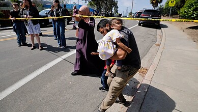 A woman carrying a child walks past a police scene with yellow tape and onlookers on the street.