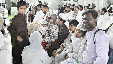 A young boy speaking to a seated group of men and children, some wearing traditional Islamic attire, in a.