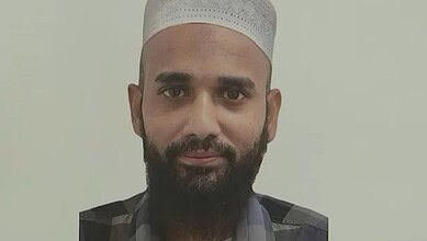 A man with a beard wearing a white cap and traditional attire, looking directly at the camera.