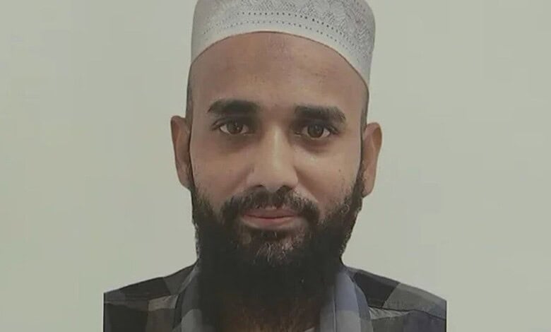 A man with a beard wearing a white cap and traditional attire, looking directly at the camera.