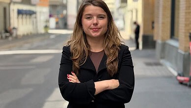 A woman with long brown hair smiling confidently on a city street in daytime.
