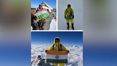 A mountaineer holding the Indian flag on a snowy peak, dressed in yellow gear with a clear blue sky backg.