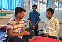 A man in a striped shirt talking to a patient in a white shirt in a hospital room, with a nurse standing.