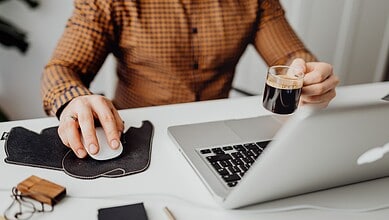 A person working on a laptop with a cup of coffee in hand at a desk.