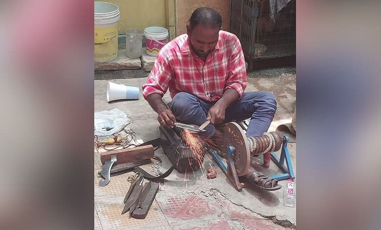 A man welding metal outdoors with sparks flying, seated on the ground, surrounded by tools and containers.