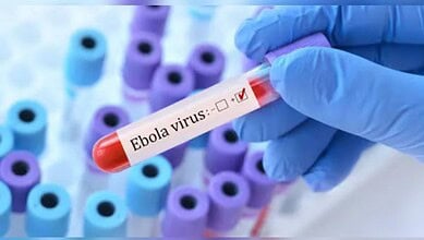 Ebola virus test tube held by gloved hand with multiple test tubes in background.