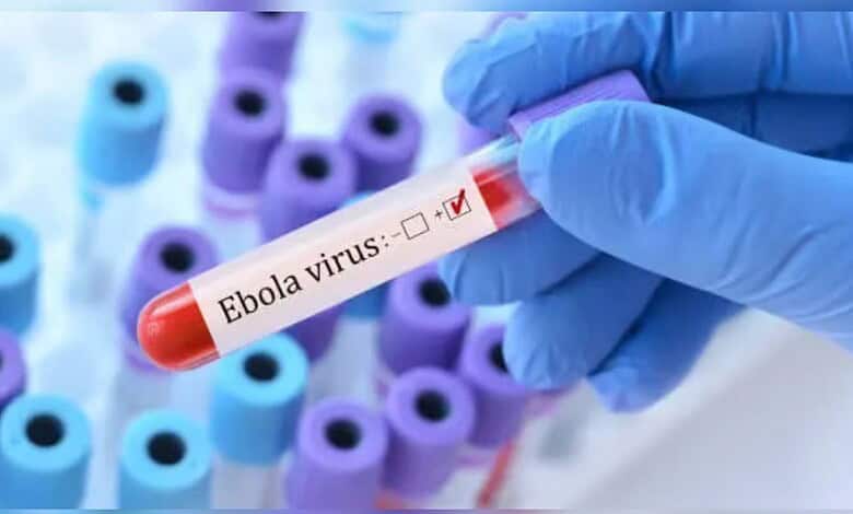 Ebola virus test tube held by gloved hand with multiple test tubes in background.
