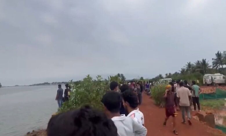 Flood-affected people gather along a riverbank in a rural area, with some standing and others walking, un.