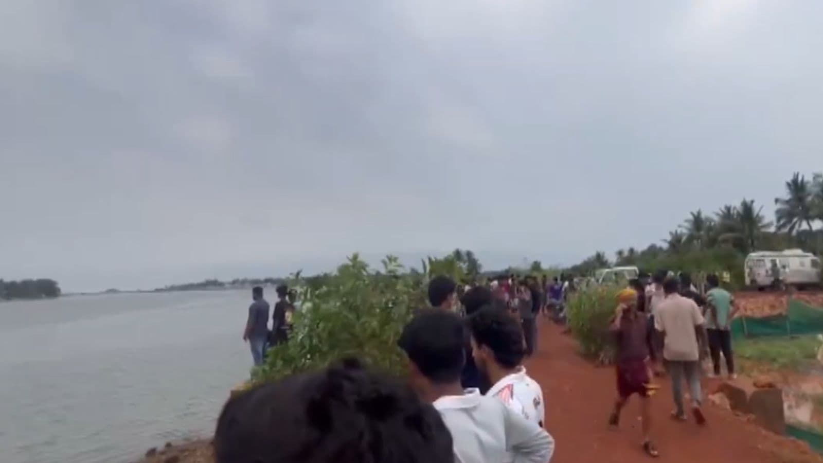 Flood-affected people gather along a riverbank in a rural area, with some standing and others walking, un.
