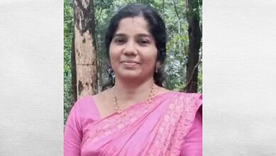 A woman in a pink saree smiling outdoors in a forest setting.