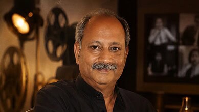 A man with a moustache and grey hair, wearing a black shirt, smiling in a warmly lit room with vintage de.
