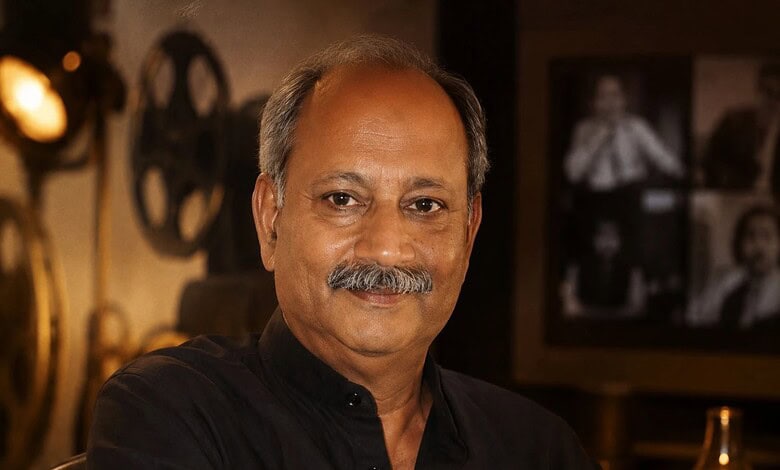 A man with a moustache and grey hair, wearing a black shirt, smiling in a warmly lit room with vintage de.
