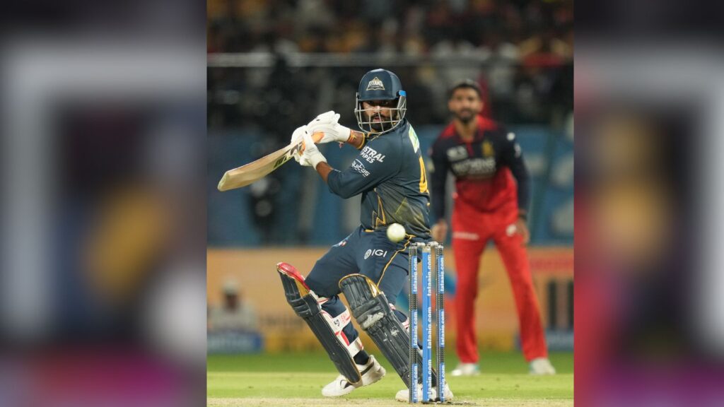 A cricket player in navy blue uniform batting during a match with a bowler in red and black in the backgr.