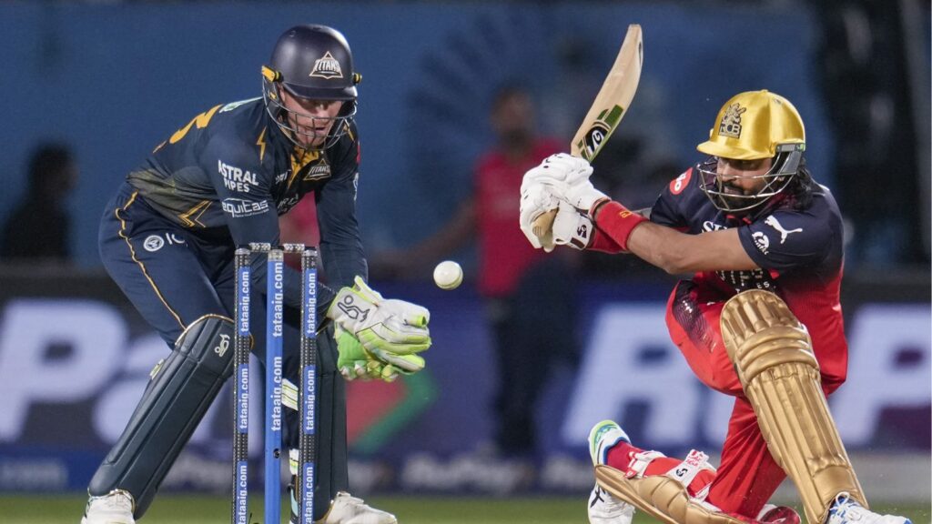 A cricket player in red and black pads hits the ball while the wicketkeeper in blue gear prepares to catc.