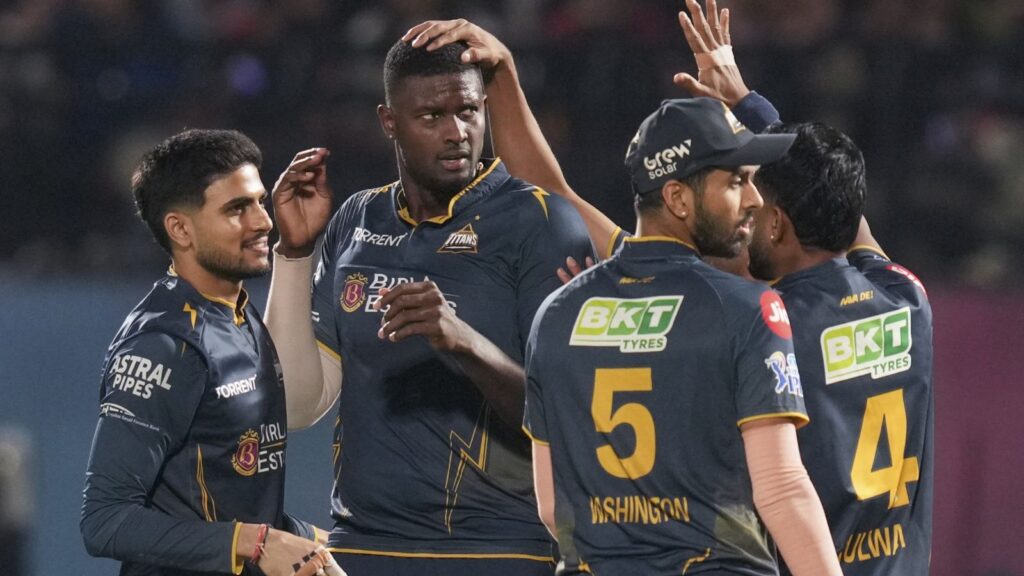 Cricket players celebrating on the field during a match, wearing dark blue uniforms with sponsor logos.