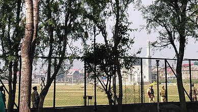 Bright outdoor sports field with players and trees, enclosed by a black fence, under a clear sky.
