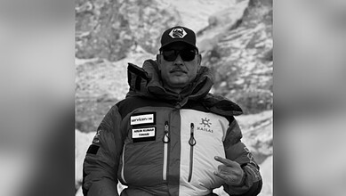 A man in outdoor gear making a peace sign in a mountainous environment, wearing sunglasses and a cap.