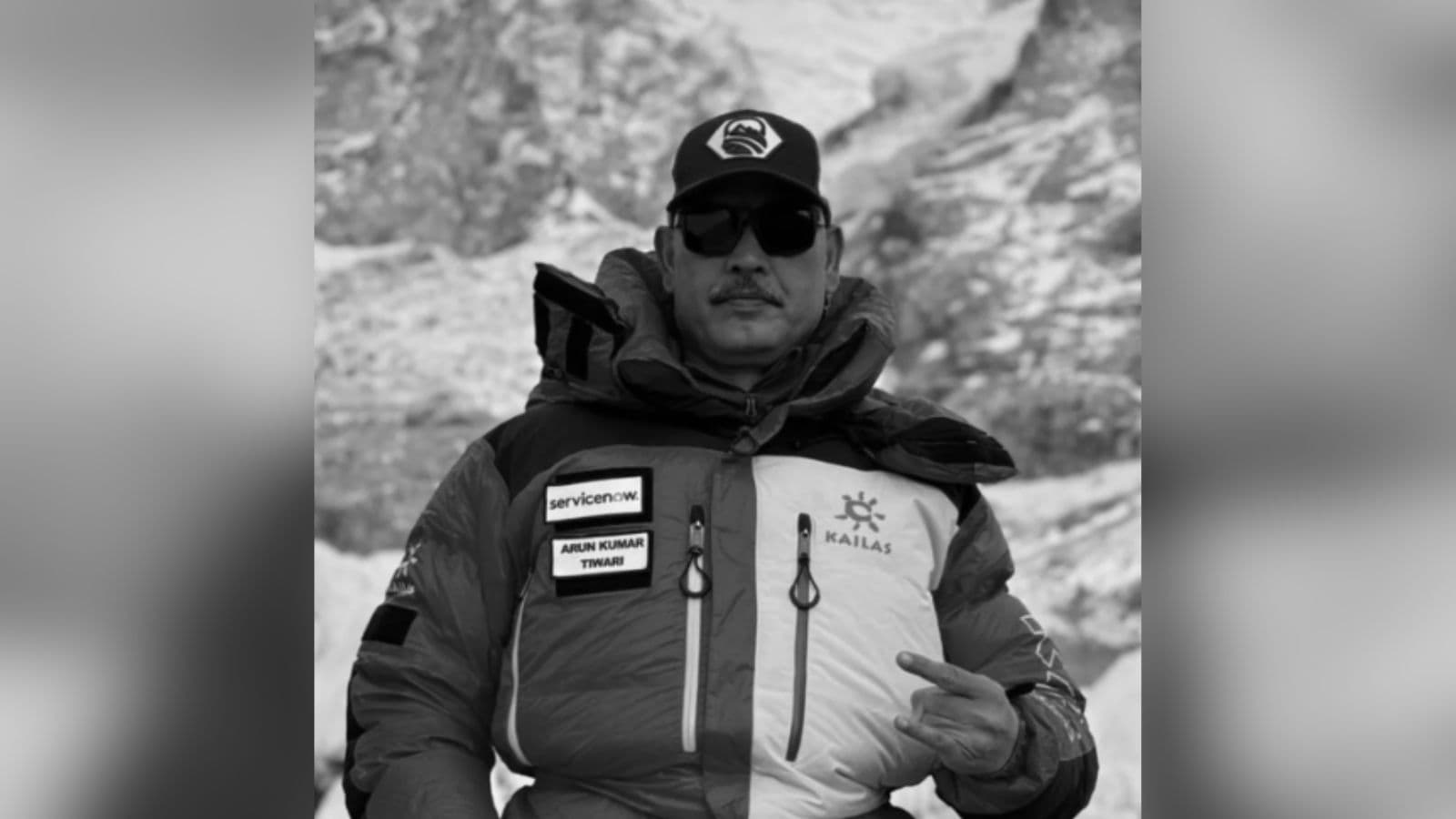 A man in outdoor gear making a peace sign in a mountainous environment, wearing sunglasses and a cap.