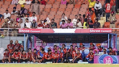 A football team sitting on the bench during a match at a stadium with spectators in the background.