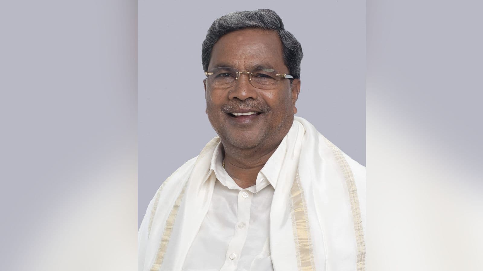 A man smiling wearing glasses and traditional white attire against a plain background.