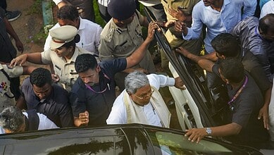 A man in white traditional attire surrounded by police and media personnel at a crowded outdoor event.