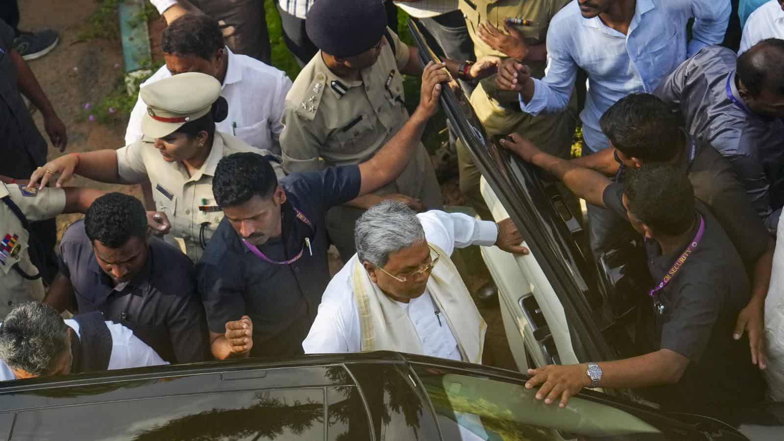 A man in white traditional attire surrounded by police and media personnel at a crowded outdoor event.