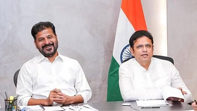 Two men in white shirts sitting at a desk with Indian flag in the background, smiling and engaged in disc.