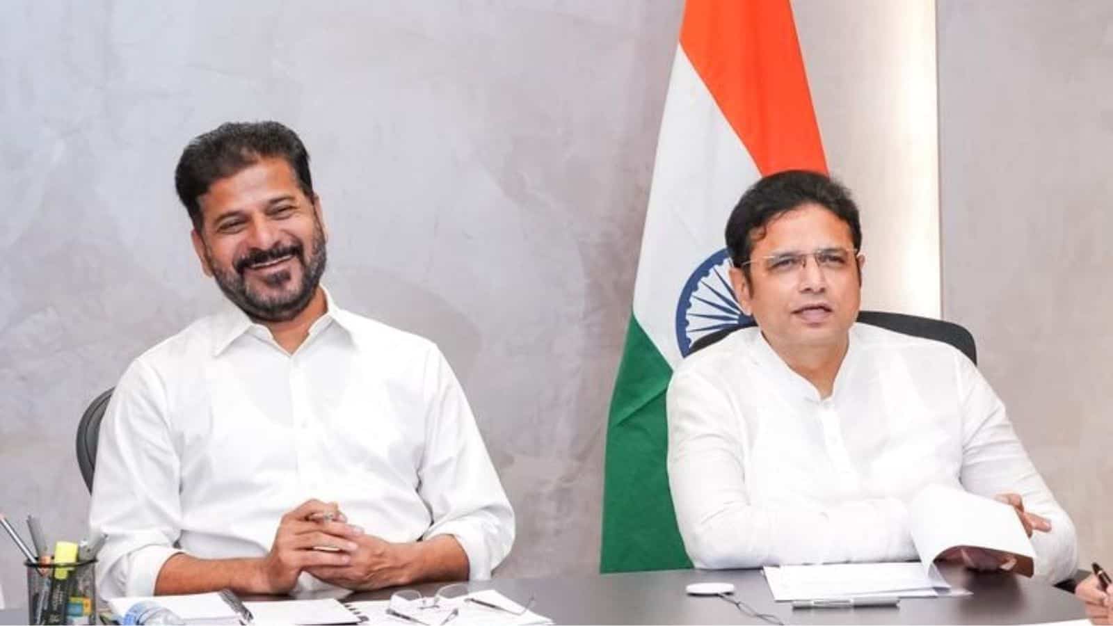 Two men in white shirts sitting at a desk with Indian flag in the background, smiling and engaged in disc.
