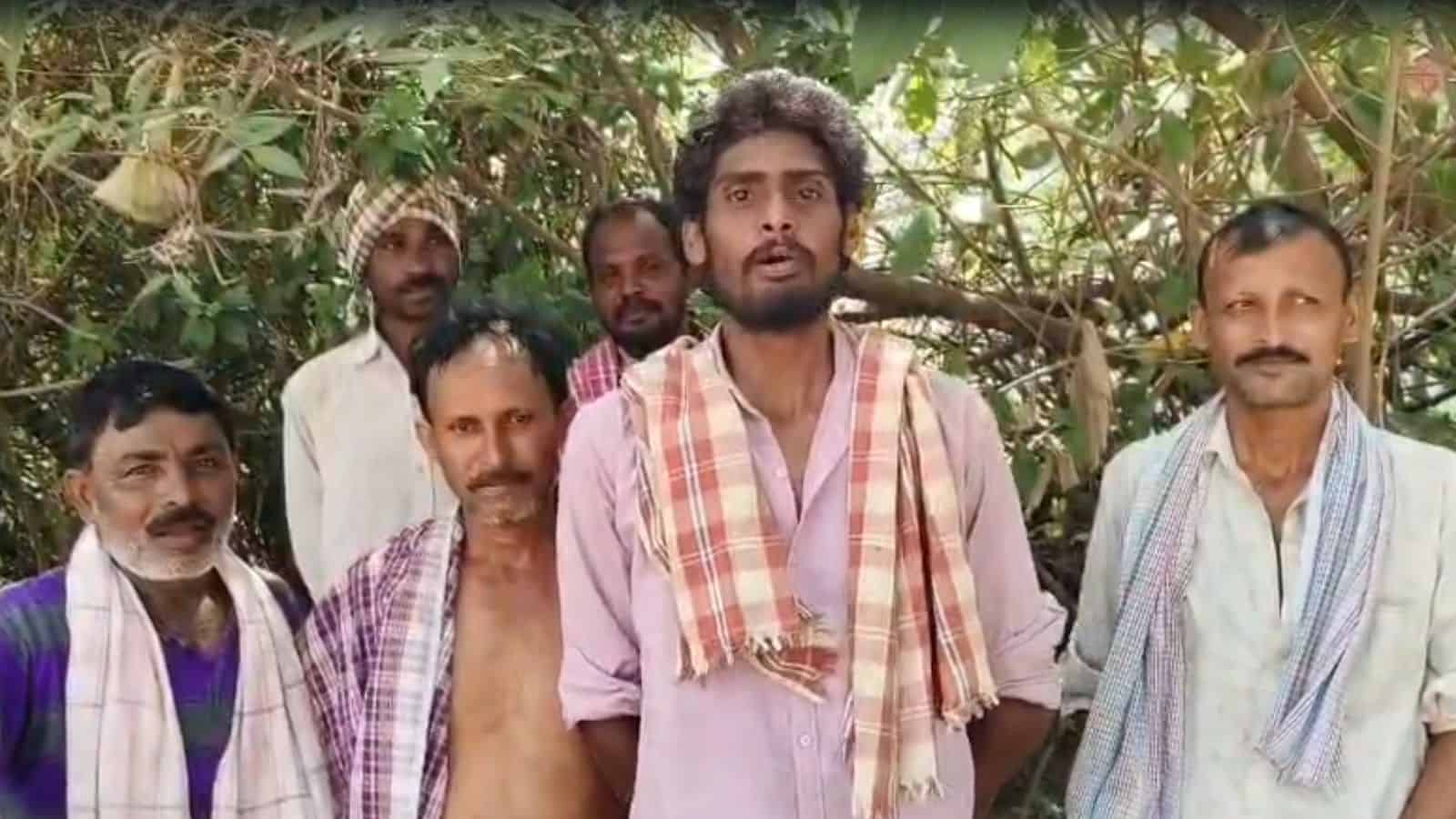 A man speaking passionately to a group of men outdoors in a rural setting with trees and foliage.